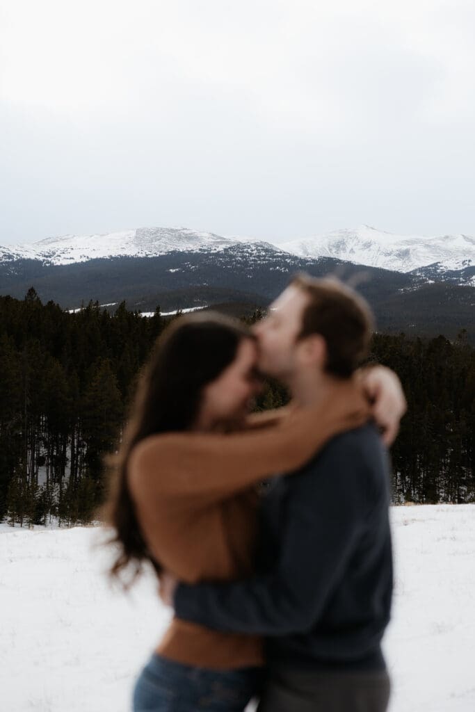 Photograph of engagement session in the Big Horn Mountains where the man is kissing the forehead of the woman, with the mountains being in focus in the background.
