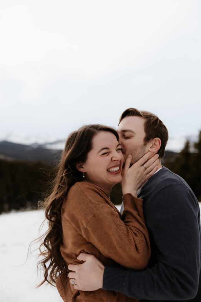 Photograph of engagement session in the Big Horn Mountains where the man is kissing the woman's cheek and she is laughing.