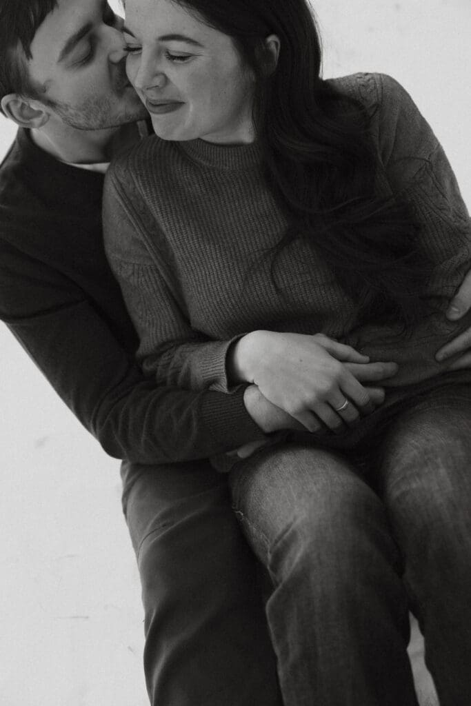 Photograph of engagement session in the Big Horn Mountains where the couple are sitting on the ground with the woman between the mans legs, and the man kissing her cheeks and she laughs. 