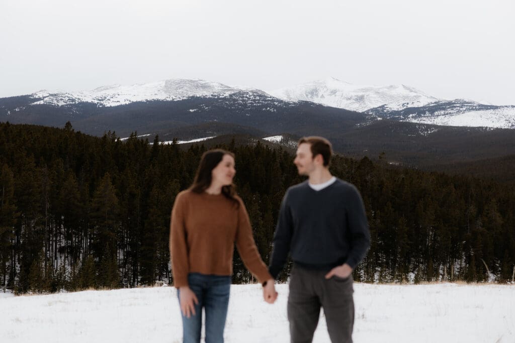 Photograph of engagement session in the Big Horn Mountains where the couple are standing side by side, holding hands, and looking at each other, with the mountains in the background being the focus.