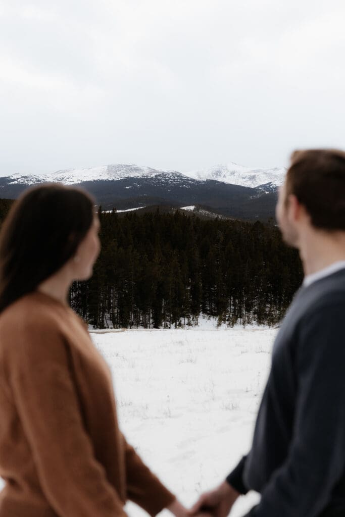 Photograph of engagement session in the Big Horn Mountains where the couple are holding hands, looking out at the Big Horn Mountains with the mountains being in focus.