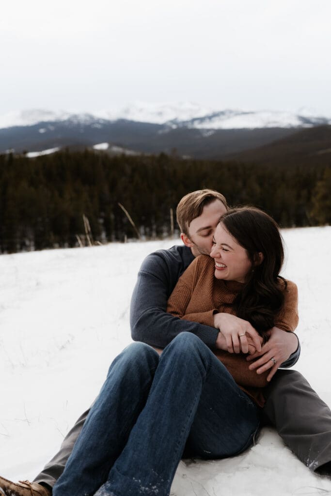 Photograph of engagement session in the Big Horn Mountains where the couple are sitting on the ground with the woman between the mans legs, and the man kissing her cheeks and she laughs. 