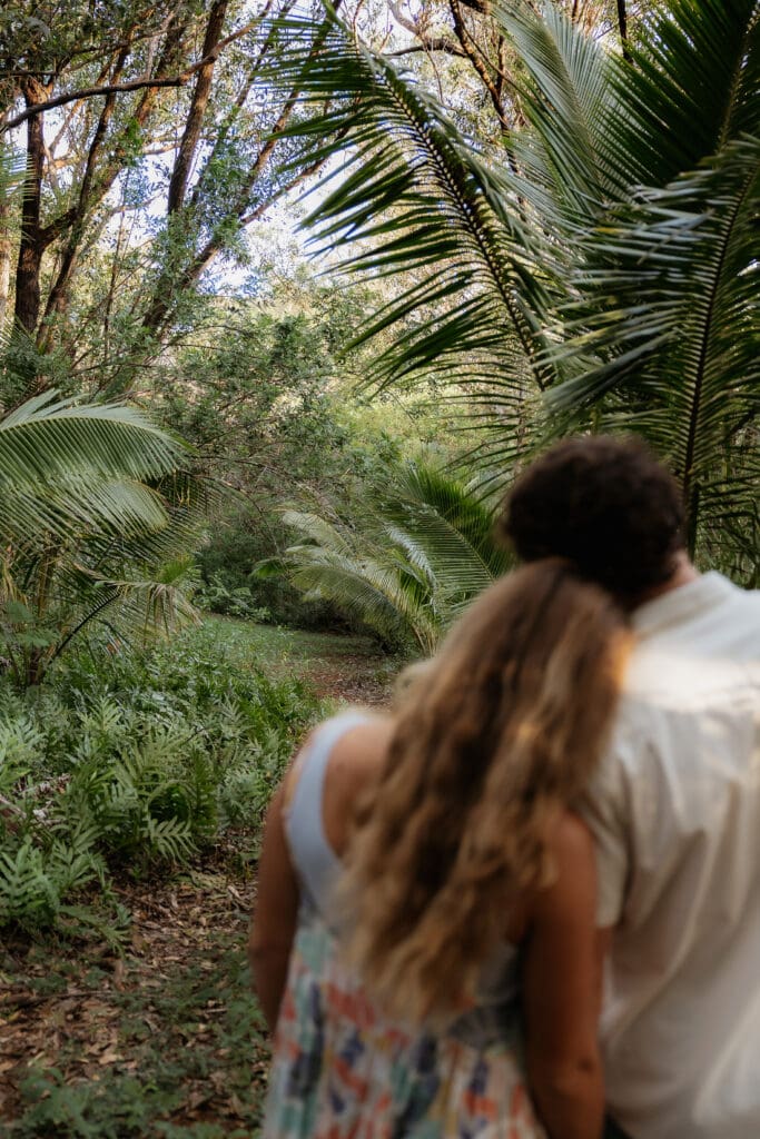 Engagement photo of man and woman standing side by side, resting their heads of each other with their backs to the camera with the jungle foliage in focus in front of them.