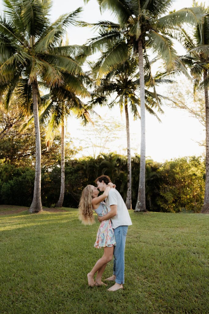 Engagement photo of man and woman facing each other and just barely about to kiss, surrounded by coconut trees and palm trees.