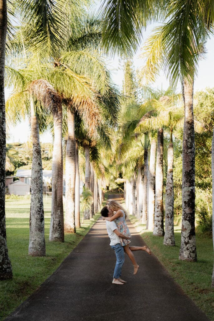 Engagement photo with man picking up woman and kissing on a long driveway surrounded by coconut trees that line the driveway. 