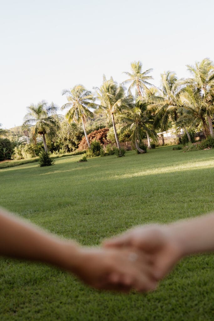 Engagement photo of man and woman holding hands with the background being in focus with coconut trees, palm trees, and a grass field.