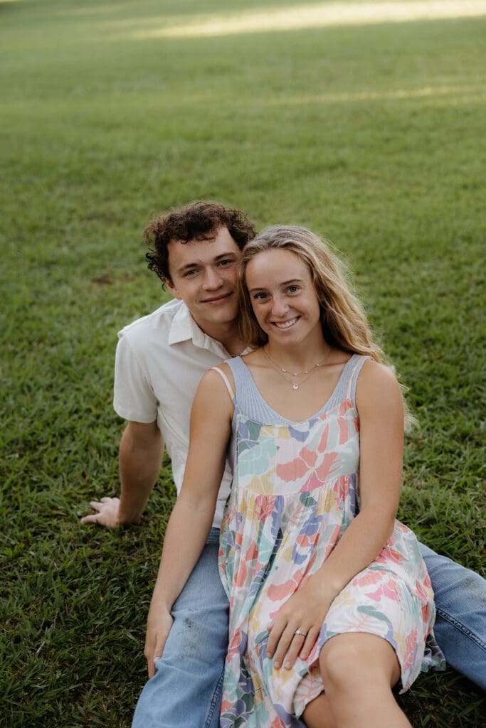 Engagement photo with woman sitting between the mans legs on a grass field and looking at the camera smiling.