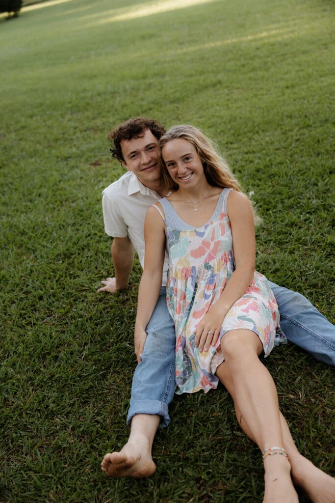 Engagement photo with woman sitting between the mans legs on a grass field and looking at the camera smiling.