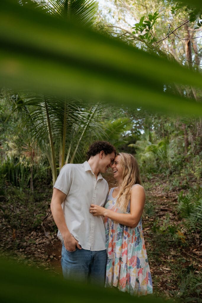 Engagement photo with jungle leaves in the foreground, man and woman standing side by side, foreheads leaned on each other while they are laughing and smiling.