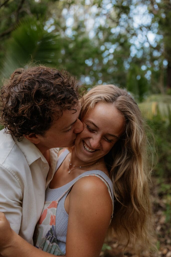 Engagement photo close up of man kissing girls cheek and she looks towards the ground and smiles.